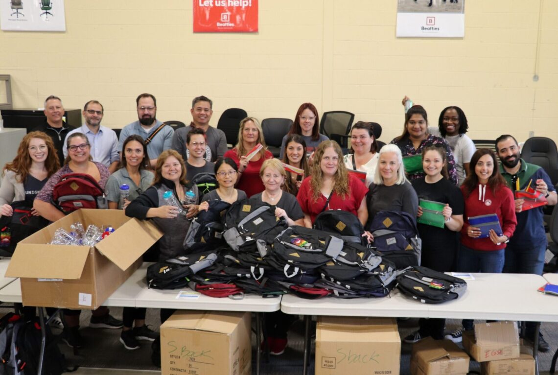 Group photo of the Algoma volunteers behind the assemby line of backpacks.